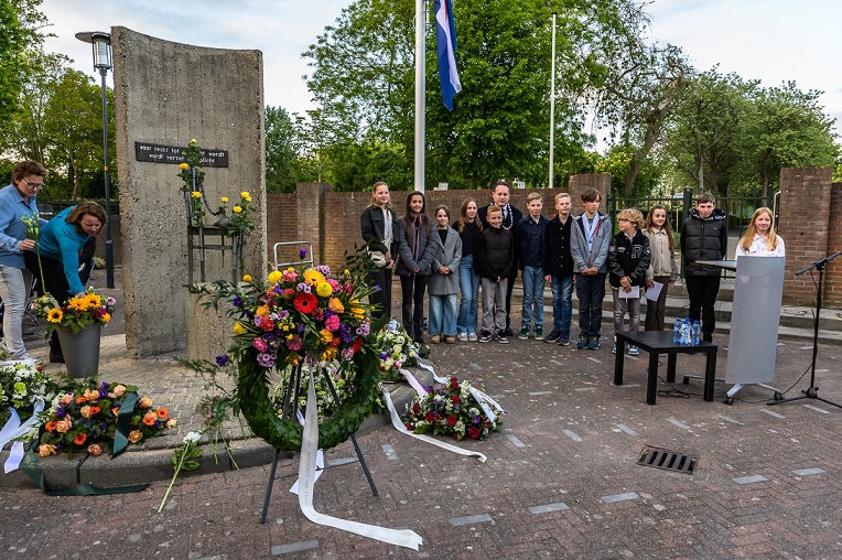Monument met kransen en daarachter burgemeester Marvin Polak met schoolkinderen die de namen van slachtoffers hebben genoemd en bloemen hebben neergelegd